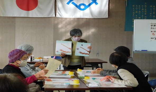 机を囲んで座る高齢者たちの前で、講師の女性が赤と黒の模様が描かれた2枚の作品を手に持ち、ちぎり絵や貼り絵の作り方を説明しているワークショップの様子の写真
