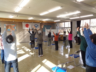 明るい室内で、たくさんの参加者たちが一列に並んだ椅子の後ろに立ち、講師の動きに合わせて一斉に両腕を高く突き上げて体操に取り組んでいる様子の写真