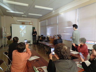 前方のスクリーンに映し出されたスマートフォンの操作画面を見ながら、着席した高齢者たちが自分のスマホを掲げて熱心に撮影や操作練習を行っている、初心者向けのスマートフォン教室の様子を写した写真