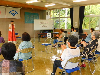 公民館のような室内で、赤いロングスカートの女性がマイクを持って前に立ち、椅子に座った大勢の高齢者が拍手をしている、口笛鑑賞講座の様子を撮影した写真