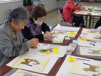 高齢の女性たちが長机に並んで座り、筆やハサミを使いながら色紙に干支のちぎり絵の作成に取り組んでいる様子の写真