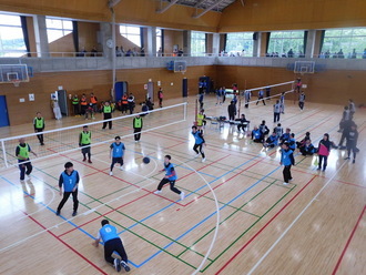 体育館のコート内で、ビブスを着用した選手たちがネットを挟んでラバーバレーボールの試合を行っている写真