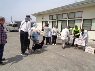 平屋の建物の前で、犬を連れた人々が一列に並び、狂犬病予防接種の受付を行っている写真