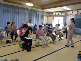畳敷きの広間で、パイプ椅子に座った高齢者を中心とする参加者たちが手元の資料を見ながら、前方に立つ女性講師の話を聞いているいきいき教室の活動の様子写真