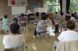 日の丸が掲げられた室内で、キーボードの前に立つ女性講師の方を向き、パイプ椅子に座った参加者たちがリズムに合わせて拍手をしている第2回宇津峰ときめきカレッジ講座の様子を後方から撮影した写真