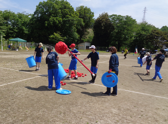 晴れ渡った空の下、小学校の校庭で青いジャージと紺色のTシャツを着用した生徒たちが、運動会の玉入れ競技で使った赤い玉を青いバケツに回収したり、背の高い籠の支柱を手に持っている写真