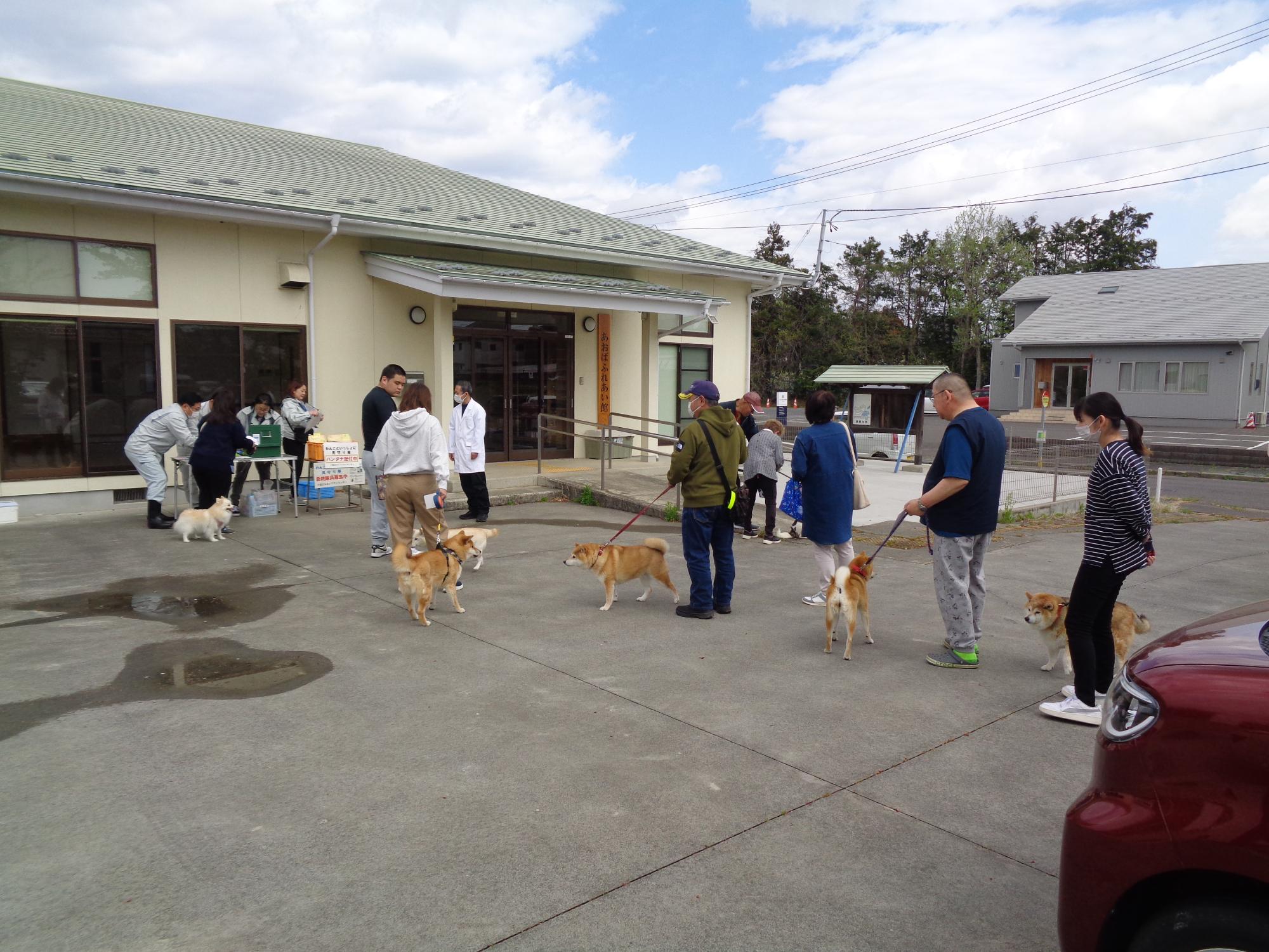 平屋の建物の前で、犬を連れた人々が一列に並び、狂犬病予防接種の受付を行っている写真