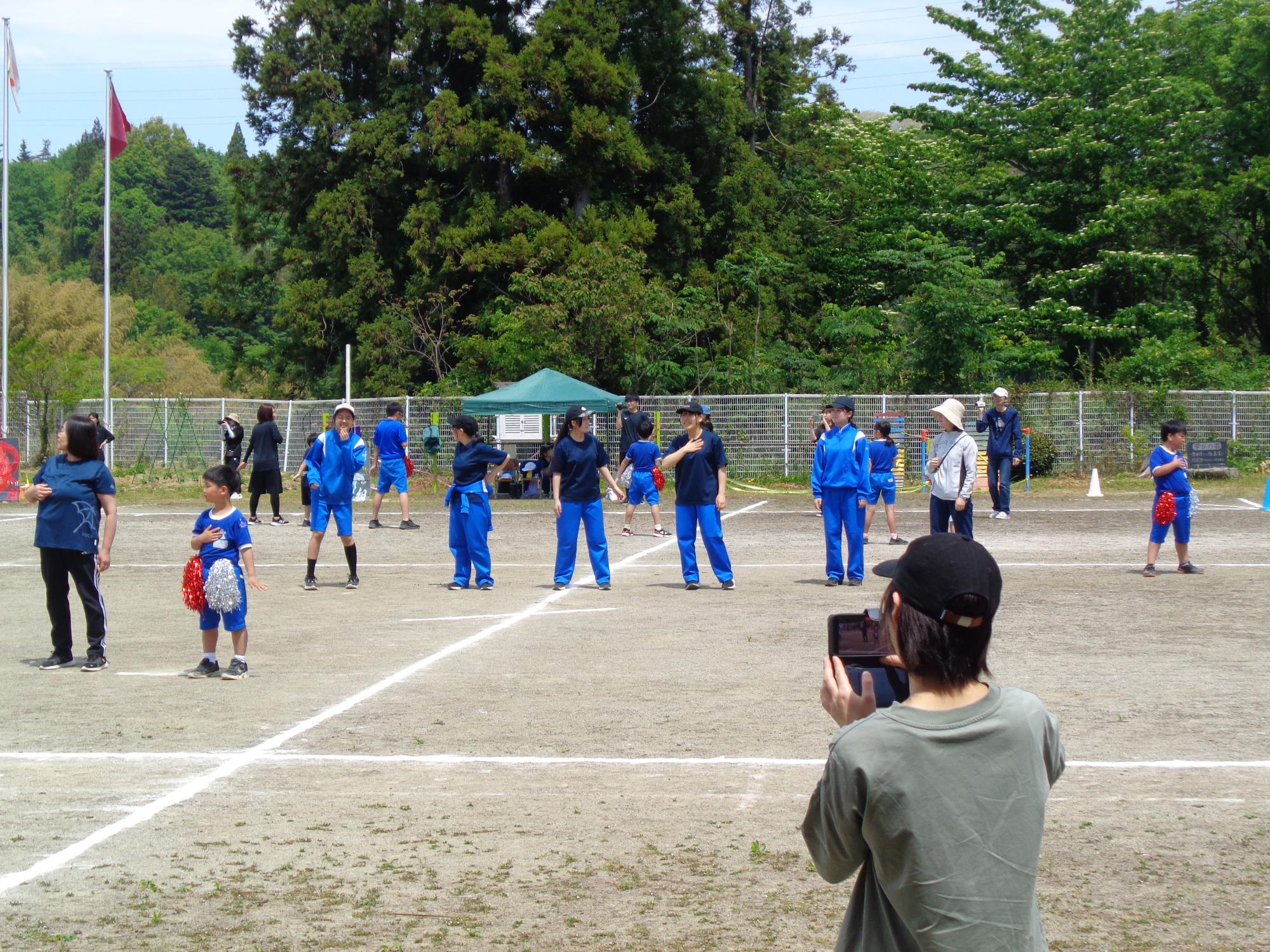 晴れ渡った空の下、小学校の校庭で青いジャージと紺色のTシャツを着用した生徒たちが、運動会のプログラムに合わせて活動している写真