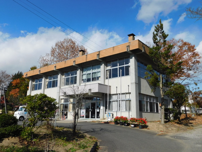 青空の下に立つ、大きな窓が並ぶコンクリート造りの2階建て施設で入り口付近に並べられた赤い花、周囲の木々やわずかに色付いた葉が秋の訪れを感じさせる写真