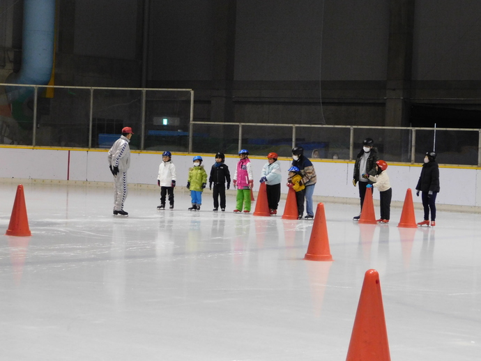 アイススケートリンクで、赤い帽子をかぶったインストラクターの前に、色とりどりの防寒着とヘルメットを着用した子どもたちが一列に並び、赤いコーンが置かれた氷上でスケート教室を受けている様子の写真