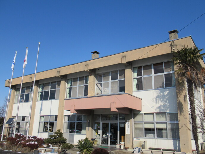 低木の植栽と3本の旗のポールが並んだ後ろに建つ2階建ての仁井田公民館の外観写真