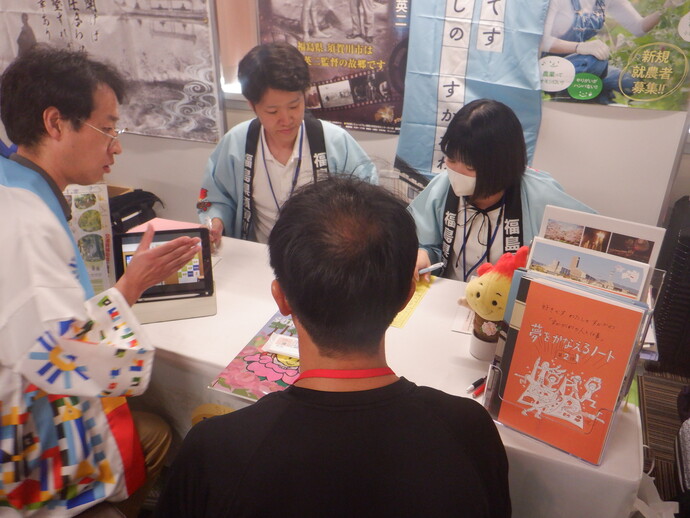 イベント会場で、法被姿の相談員がタブレット端末などを用いて、ブースを訪れた男性に福島県須賀川市での生活や新規就農について熱心に説明を行っている写真