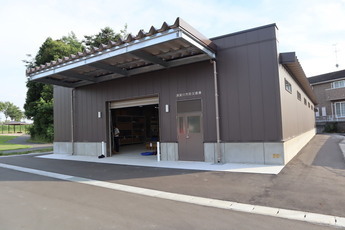 茶色の外壁と金属製の庇を持つ平屋建ての建物が、屋外の舗装された広場に建っており、中央のシャッターが開いている倉庫の外観写真