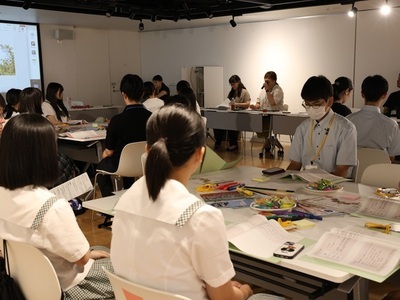 複数の学生が資料や文房具を広げた机を囲んで話し合い、前方では発表を聞きながら活動内容を発表している様子の写真