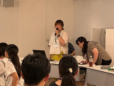 会場前方で白い服を着た女性がマイクを持って説明し隣のスタッフが準備を支え参加者が着席して聞き入る様子の写真