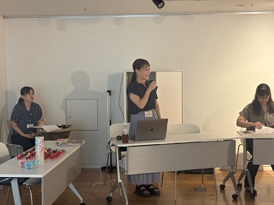 ノートパソコンを前にマイクを握ってにこやかに話す女性と、その左右で資料の確認や進行を見守るスタッフたちの様子を捉えた写真