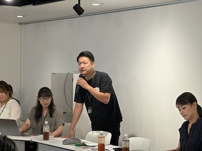 マイクを手に立ち上がって力強く話す男性と、その横でノートパソコンを操作したり資料を確認したりしながらサポートするスタッフたちの様子を捉えた写真