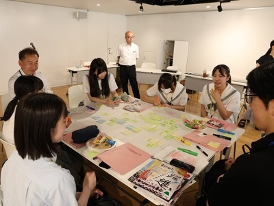 数人の若者や大人がテーブルを囲み、付箋が貼られた模造紙や資料を広げながら真剣な表情で話し合いや作業に取り組んでいる様子の写真