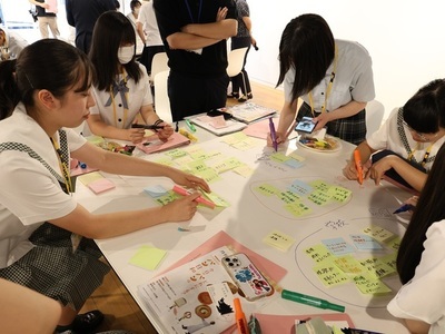 学生たちが机いっぱいに並べた付箋や資料へ丁寧に書き込みながら意見を整理し協働して作業を進めている写真