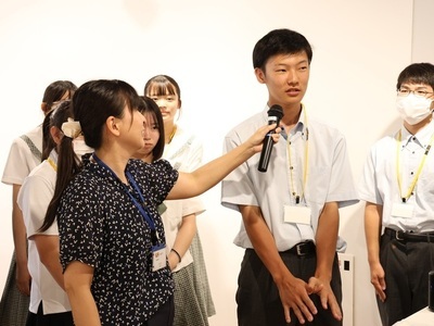 マイクを向けられた男子学生が緊張した面持ちで話し、その傍らで女子学生や別の男子学生が静かに見守っている様子の写真