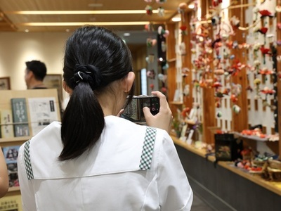 店内で女子生徒がカメラを構えて色鮮やかな吊るし飾りを撮影している様子を背後から撮影した写真