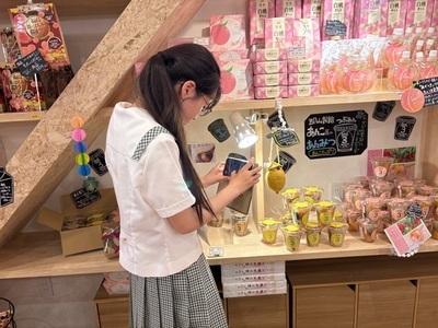 店内で女子生徒が棚に並ぶ桃を使った菓子や土産物を手に取り撮影し商品陳列の様子を記録している写真