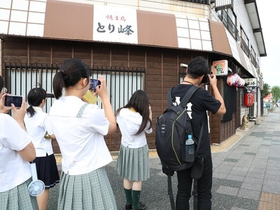 商店街の通りで学生が店先の看板や建物外観にカメラを向けて撮影している様子の写真