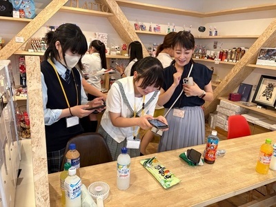 店内のテーブルで学生たちが飲み物や文具の並ぶ作業スペースを囲み、スマートフォンを使って協力しながら情報を確認している写真
