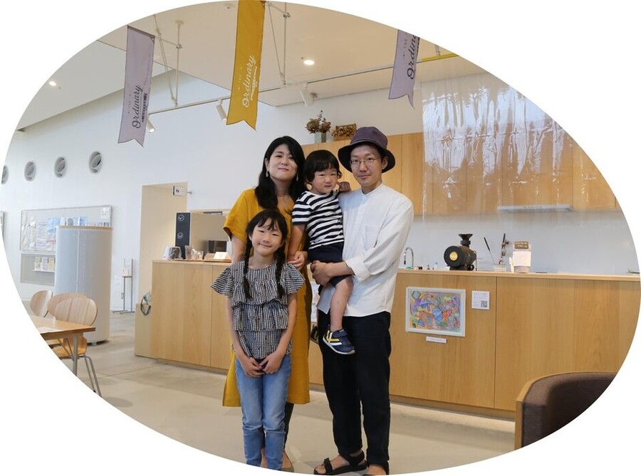 カフェ風の明るい室内で、木製カウンターの前に斎藤さん家族4人が立ち、カウンターにはコーヒーメーカーや装飾品が置かれ、壁には絵画と透明なカーテンが掛けられ、天井からは「Quality」などの文字が入った黄色と白のバナーが吊り下がり、左側には椅子とテーブル、奥には冷蔵庫が見える写真