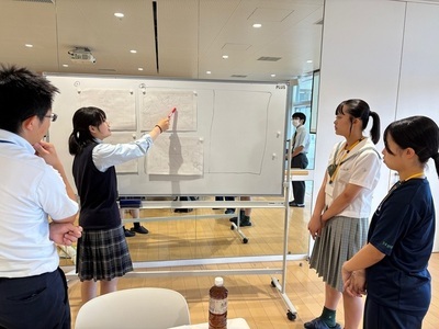 女子学生がホワイトボード上の資料を指し示しながら周囲の仲間に説明し、意見を交わしつつ学習内容を共有している写真
