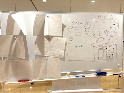 ホワイトボードに複数の紙資料が貼られ、右側にはタイトルや配置案などの手描きレイアウト図が詳細に示されている写真