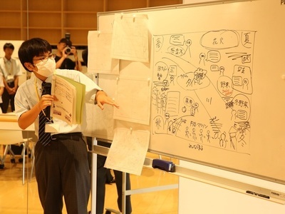 男子学生がホワイトボードに描かれたレイアウト図を指し示し、手元の資料を用いて内容を説明している写真