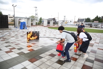 広場で火災訓練が行われており、大人が子供の手を取りながら協力して消火器を操作し、燃え上がる模擬火災に向けて水を噴射している様子の写真