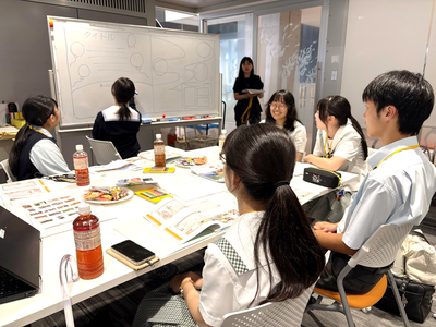 室内で学生たちが資料の置かれた机を囲んで着席し、発表者が示すホワイトボードの図や説明に集中して耳を傾けている写真