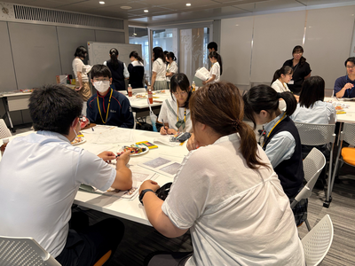 室内で複数の学生が資料を囲んで意見交換や作業を進め、周囲でも別グループが協働的に活動している写真