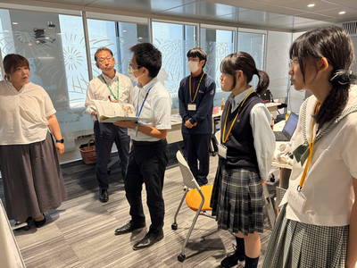 室内で指導者が資料を手に説明し学生たちが真剣に耳を傾けながら周囲に集まって活動内容を確認している様子を捉えた写真