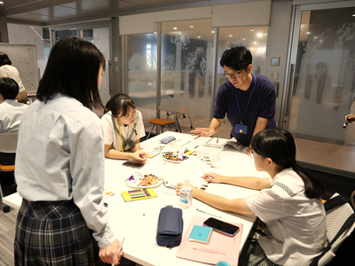 室内で女子生徒3人が机を囲んで資料や文具を使いながら指導者の助言を受け、落ち着いた雰囲気の中で共同作業を進めている写真