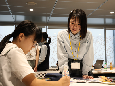 室内で女生徒が机上の資料に向かって作業し、その横で女性が立って内容を確認しながら協力して課題に取り組む様子の写真