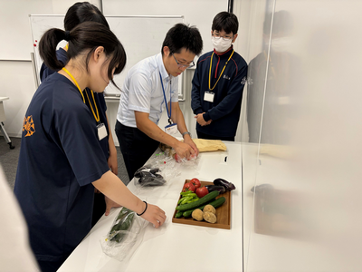室内で指導者と学生がテーブルに並べたナスやトマトなどの野菜を使って作業手順を丁寧に確認しながら準備に取り組む様子の写真