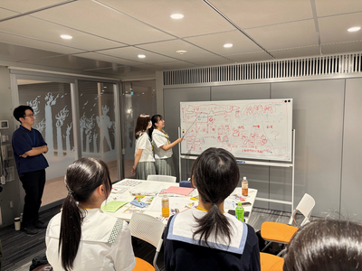 2人の女子生徒が詳細な構成図の描かれたホワイトボードを指し示し発表し周囲の仲間が着席して説明に集中して耳を傾けている写真