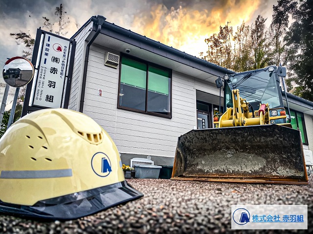地面に置いた黄色いヘルメットとショベルカーのショベル越しに、グレーとオレンジ色が混ざった幻想的な空を背景に建つ、「赤羽組」の看板が付いた薄いグレーの外壁の社屋を撮影した写真