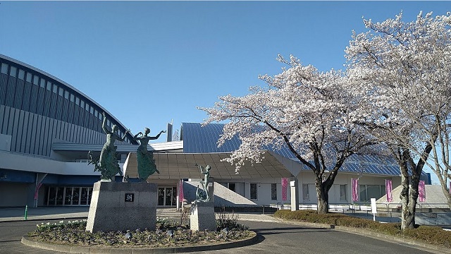 モダン的なデザインの大きなアリーナ施設の前に、満開の桜の木が植えられた植栽があり、入口正面にある円型の植栽の中央には、躍動感のある2体の女性像と、ラッパを吹く人物の像の2つのモニュメント並んでいる様子の写真