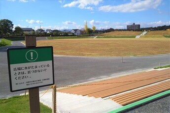 舗装された道路に面した広大な茶色い広場があり、手前には注意書きが書かれた緑の看板が設置されている公園の写真