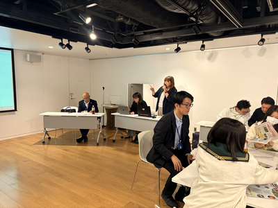室内で黒のジャケットを着た女性が立って指示を出し前方の机でスタッフが機材を操作し参加者が資料を広げて話し合いながら作業を進める様子の写真