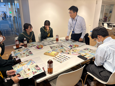 テーブルを囲む学生たちが配布資料を見ながら意見を交わし、立っている男性の指導者が内容を示して作業を支援している写真