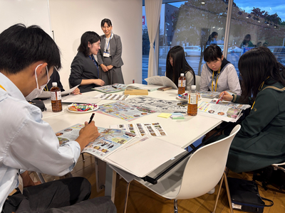 学生たちが資料を広げた机を囲んで作業に集中し、女性の指導者がそばで進行を見守りながら助言する様子を撮影した写真