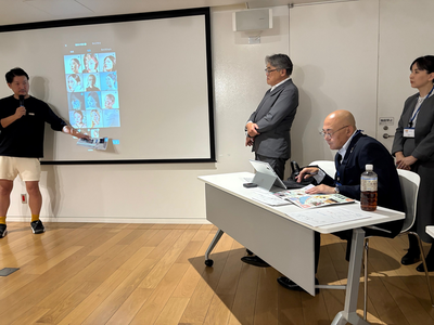 男性の発表者が前方のスクリーンに人物イラストが並んだスライドを映し出して説明し、机側の参加者が資料を確認しながら聞き、スーツ姿の男性が進行を見守っている写真