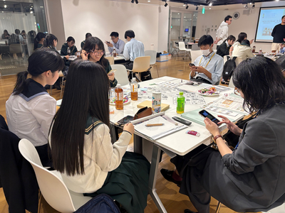 会議室で5人の学生が資料を広げた机を囲みスマートフォンや文具を使って情報を確認しながら意見交換と作業を進めている写真