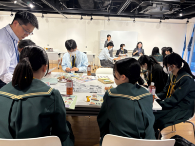 室内で5人の学生たちと男性の指導者が資料を広げた机を囲んで意見を書き込みながら協力して作業を進めている写真