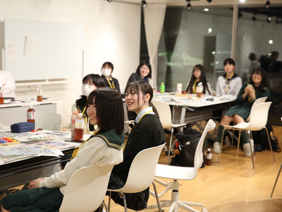 学生たちが机を囲んで着席し前方の発表に耳を傾けながら互いに笑顔を交えつつ活動の様子を見守っている写真